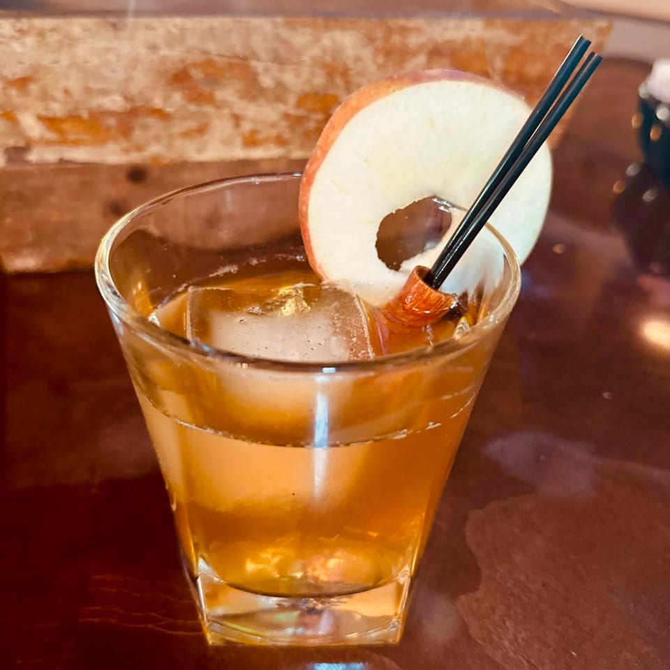 Whiskey cocktail with ice, an apple slice, and a cinnamon stick on a wooden table.
