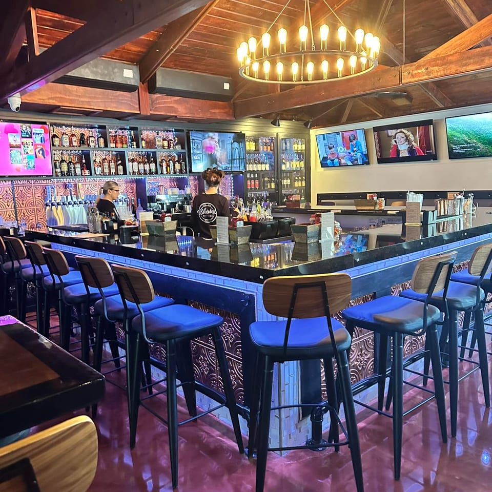 Whiskey Kitchen bar with blue stools, liquor bottles, TVs, and two bartenders working.