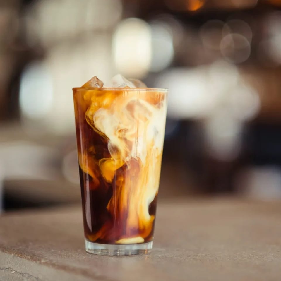 Iced coffee with cream swirling into dark liquid, served in a tall glass on a counter.