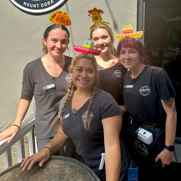 Four smiling Whiskey Kitchen staff members wearing festive taco and sombrero headbands outdoors.