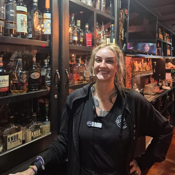 A smiling bartender stands in front of a well-stocked bar with various liquor bottles and beer taps.