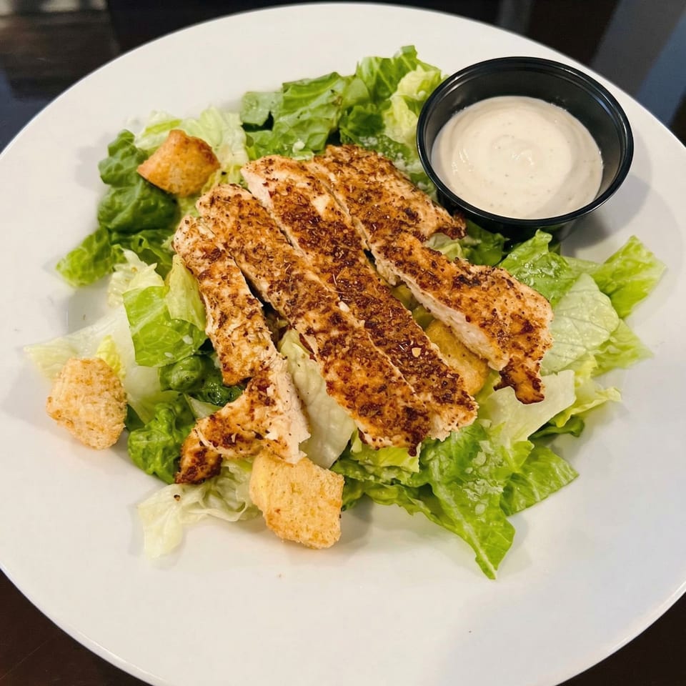 Grilled chicken Caesar salad with croutons and a side of dressing on a white plate.